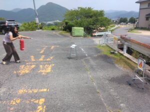 屋外で女性研修生が赤い消火器のホースを持ち、火の絵が描かれた的に向けて水を噴射している様子